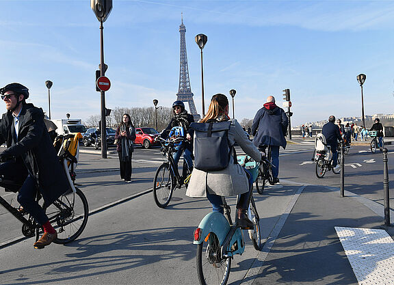 Menschen auf Fahrrädern überqueren eine Straße in Paris. Im Hintergrund ist der Eiffelturm zu sehen.